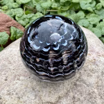Handcrafted glass cremation paperweight shown from a top view, featuring black, white, and blue rippled swirl patterns with encased cremation ashes throughout the piece, displayed outdoors on a natural stone surface with garden greenery in the background