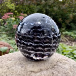 Handcrafted glass cremation paperweight with a rippled black and white swirl design containing encased cremation ashes, displayed outdoors on a natural stone with garden greenery in the background to highlight color, texture, and form