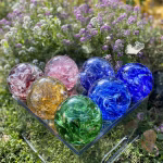 Group view of seven Spirit Orb cremation paperweights in assorted colors—red, gold, green, pink, light blue, cobalt blue, and deep blue—crafted from swirling glass mixed with cremation ashes. The handcrafted memorial orbs are arranged on a clear display box outdoors, surrounded by blooming purple and white flowers and sunlight, creating a vibrant remembrance setting.