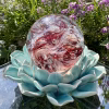 Angled front view of a Red Spirit Orb cremation paperweight, a hand-blown glass memorial sphere with swirling red and white patterns infused with cremation ash, displayed on a turquoise ceramic lotus stand outdoors with sunlight and blooming purple garden flowers in the background.