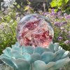 Front-view of a Red Spirit Orb cremation paperweight, a hand-blown glass memorial sphere with swirling ruby and clear patterns and cremation ash inclusions, resting on a turquoise ceramic lotus stand outdoors with bright sunlight and blooming purple and white garden flowers in the background.