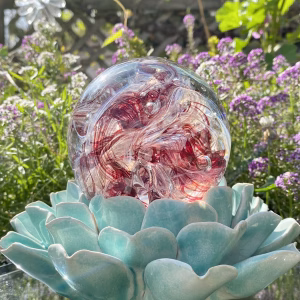 Front-view of a Red Spirit Orb cremation paperweight, a hand-blown glass memorial sphere with swirling ruby and clear patterns and cremation ash inclusions, resting on a turquoise ceramic lotus stand outdoors with bright sunlight and blooming purple and white garden flowers in the background.