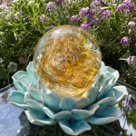 Front-view of a Golden Spirit Orb cremation paperweight, a hand-blown glass memorial keepsake with swirling amber tones and cremation ash inclusions, displayed on a turquoise ceramic lotus stand outdoors against a vibrant garden backdrop of blooming purple and white flowers.