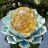 Top-view of a Golden Spirit Orb cremation paperweight, a handcrafted glass memorial keepsake infused with cremation ashes, displayed on a turquoise ceramic lotus stand outdoors with blooming garden flowers and greenery surrounding it.