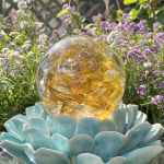 Golden Spirit Orb cremation paperweight, a handcrafted glass memorial keepsake infused with cremation ashes, shown in a front garden-view display resting on a turquoise ceramic lotus stand with blooming purple and white flowers in the background.