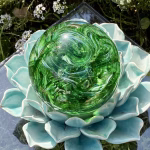 Top-down view of a green Spirit Orb cremation paperweight, a handcrafted swirling glass memorial infused with cremation ashes, resting on a turquoise ceramic lotus stand outdoors with blooming white flowers and greenery surrounding it.