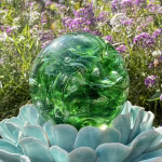 Close-up view of a green Spirit Orb cremation paperweight, a handcrafted swirling glass memorial keepsake infused with cremation ashes, displayed outdoors on a turquoise ceramic lotus stand with purple flowers and greenery in the background.