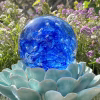Front view of a blue Spirit Orb cremation paperweight featuring swirling cobalt and white glass with ashes encased inside. The handcrafted memorial orb is displayed on a turquoise ceramic lotus and photographed outdoors with blooming purple flowers in the background, creating a peaceful remembrance scene.