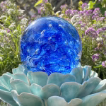 Front view of a blue Spirit Orb cremation paperweight featuring swirling cobalt and white glass with ashes encased inside. The handcrafted memorial orb is displayed on a turquoise ceramic lotus and photographed outdoors with blooming purple flowers in the background, creating a peaceful remembrance scene.
