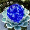 Top view of a blue Spirit Orb cremation paperweight with swirling cobalt and white glass encasing cremation ashes. The handcrafted memorial orb rests on a turquoise ceramic lotus and is photographed outdoors with greenery and small white flowers, creating a serene remembrance display.