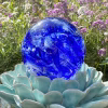 Front view of a blue Spirit Orb cremation paperweight featuring swirling cobalt and white glass with ashes encased inside. The handcrafted memorial orb is displayed on a turquoise ceramic lotus and photographed outdoors with blooming purple flowers in the background, creating a peaceful remembrance scene.