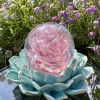 Front view of a pink Spirit Orb cremation paperweight featuring swirling rose-pink glass and delicate ashes encased within. The handcrafted memorial orb rests on a turquoise ceramic lotus and is photographed outdoors among blooming purple and white flowers, creating a peaceful and uplifting remembrance scene.