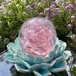 Front view of a pink Spirit Orb cremation paperweight featuring swirling rose-pink glass and delicate ashes encased within. The handcrafted memorial orb rests on a turquoise ceramic lotus and is photographed outdoors among blooming purple and white flowers, creating a peaceful and uplifting remembrance scene.