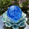 Top view of a handcrafted blue Spirit Orb cremation paperweight, showing swirling cobalt and aqua glass patterns with encased ashes throughout. The memorial orb rests on a turquoise ceramic lotus in an outdoor garden setting, highlighting its luminous color and detailed remembrance design.