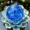 Top view of a handcrafted blue Spirit Orb cremation paperweight, showing swirling cobalt and aqua glass patterns with encased ashes throughout. The memorial orb rests on a turquoise ceramic lotus in an outdoor garden setting, highlighting its luminous color and detailed remembrance design.