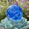 A handcrafted blue Spirit Orb cremation paperweight made from molten glass, featuring swirling cobalt and white patterns with a loved one’s ashes encased as delicate white flecks. The memorial orb rests on a turquoise ceramic lotus in bright outdoor light, creating a vibrant and serene remembrance display.
