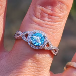 Close-up of the Infinity Cremation Ring worn on a hand. The ring features a vibrant blue cremation stone with a blue silver crinkle-glass effect, set in a round halo of small cubic zirconia. The band forms an infinity shape on both sides, accented with additional stones, shown in bright outdoor sunlight.