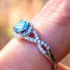 Close-up side profile of an Infinity Cremation Ring featuring a shimmering blue cremation stone set in a raised prong halo. The band forms an elegant infinity shape adorned with small sparkling cubic zirconia, shown on a finger with a softly blurred warm background.