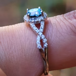 Close-up side view of an Infinity Cremation Ring on a finger, featuring a blue memorial stone set in a prong halo with sparkling cubic zirconia. The band forms an elegant infinity loop lined with stones, with a blurred natural background highlighting the ring’s detail.