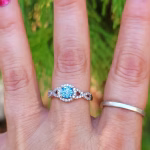Front view of an Infinity Cremation Ring worn on a hand. The ring features a brilliant blue cremation stone set in a round halo of sparkling cubic zirconia. The band forms an elegant infinity shape on both sides, also lined with small CZ stones. Shown against a soft green outdoor background.