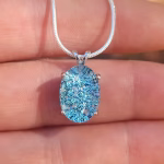Oval halo cremation pendant in sterling silver with blue silver crinkle glass, shown on chain resting on fingers.