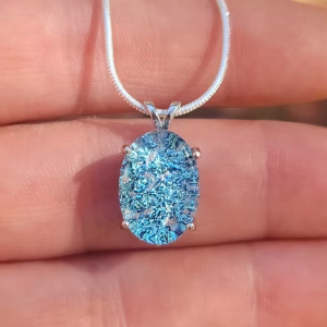 Oval halo cremation pendant in sterling silver with blue silver crinkle glass, shown on chain resting on fingers.