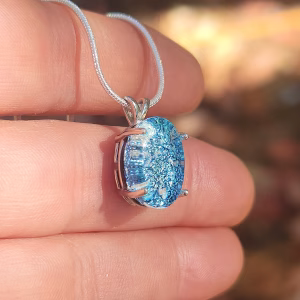 Side view of oval halo cremation pendant in sterling silver with blue silver crinkle glass, shown on chain against fingers.