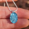Oval halo cremation pendant in sterling silver with blue silver crinkle glass, shown on chain resting on fingers.