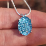 Oval halo cremation pendant in sterling silver with blue silver crinkle glass, shown on chain resting on fingers.
