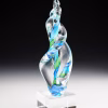 Hand-blown glass Legacy Flame memorial sculpture in swirling green and aqua tones, infused with cremation ash and displayed on a clear pedestal block, symbolizing remembrance and celebration of life.