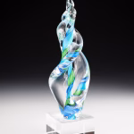 Hand-blown glass Legacy Flame memorial sculpture in swirling green and aqua tones, infused with cremation ash and displayed on a clear pedestal block, symbolizing remembrance and celebration of life.