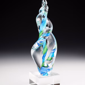 Hand-blown glass Legacy Flame memorial sculpture in swirling green and aqua tones, infused with cremation ash and displayed on a clear pedestal block, symbolizing remembrance and celebration of life.
