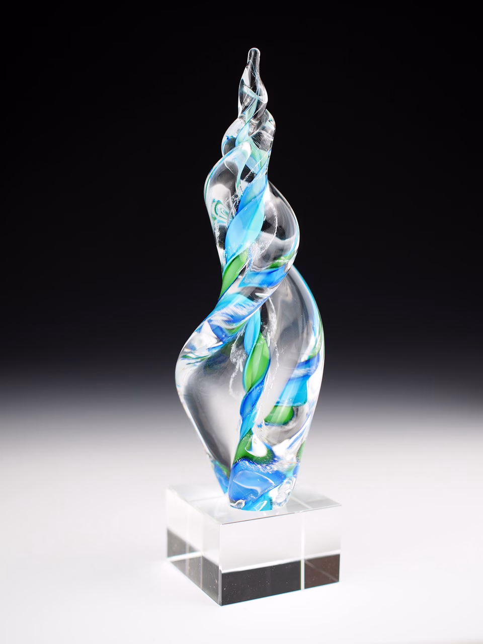 Hand-blown glass Legacy Flame memorial sculpture in swirling green and aqua tones, infused with cremation ash and displayed on a clear pedestal block, symbolizing remembrance and celebration of life.
