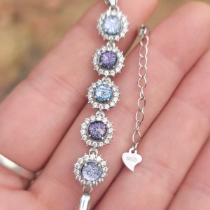 Close-up of the Dichroic Halo Cremation Bracelet resting on a hand, showing five round blue and purple dichroic memorial glass stones surrounded by clear cubic zirconia halos, set in sterling silver with an adjustable extender chain and S925 heart tag.