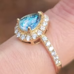 A close-up of a 14k gold cremation ring featuring a pear-shaped pale blue cremation stone framed by a halo of sparkling moissanite diamonds. More moissanite stones line the gold band, and the ring is shown worn on a finger in soft natural light to highlight its brilliance and delicate detailing.
