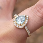 A close-up of a 14k gold cremation ring featuring a pear-shaped pale blue cremation stone framed by a halo of sparkling moissanite diamonds. More moissanite stones line the gold band, and the ring is shown worn on a finger in soft natural light to highlight its brilliance and delicate detailing.