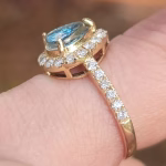 A close-up side view of a gold cremation ring featuring an oval aqua-colored center stone set in a raised halo of small round diamonds. The slim gold band is accented with a row of diamonds, and the ring is shown worn on a finger, highlighting the intricate prong and gallery detailing.