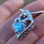 Angled side view of a mother and child cremation pendant in sterling silver, with the heart shape formed by a mother embracing her child and a blue heart-shaped memorial stone set at the center, shown on a necklace resting on a hand.