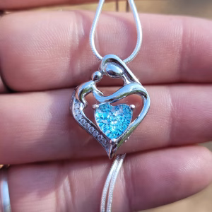 Mother and child cremation pendant in sterling silver, with the heart shape formed by a mother embracing her child and a blue heart-shaped memorial stone at the center, shown close up on a necklace resting on a hand.