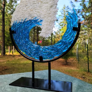 A blue and white glass ocean wave cremation sculpture displayed on the tall 10-inch black metal stand. The sculpture features textured translucent blue glass forming the body of the wave and opaque white glass creating the crest. The stand lifts the piece higher, giving it a prominent upright presentation. The background shows an outdoor forest setting with pine trees and soft natural light.