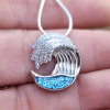 Close-up of the Ocean Wave Cremation Pendant held in hand, featuring a sterling silver wave design accented with clear cubic zirconia and a blue memorial glass inlay containing cremation ash.