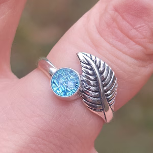 Close-up of a sterling silver leaf cremation ring featuring a round blue silver crinkle memorial ash stone, shown worn on a finger against a soft outdoor background.