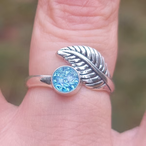 Close-up of a sterling silver leaf cremation ring featuring a blue silver crinkle memorial glass stone infused with ashes, worn on a finger outdoors.