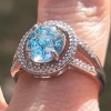 Close-up of an oval cremation ring in sterling silver featuring a bright blue memorial glass center stone with visible cremation ash, surrounded by 52 cubic zirconia accents, worn on a finger outdoors.
