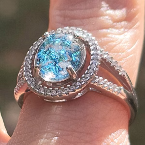 Close-up of an oval cremation ring in sterling silver featuring a bright blue memorial glass center stone with visible cremation ash, surrounded by 52 cubic zirconia accents, worn on a finger outdoors.