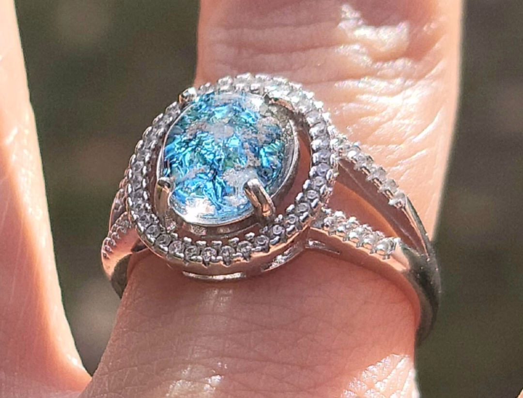 Close-up of an oval cremation ring in sterling silver featuring a bright blue memorial glass center stone with visible cremation ash, surrounded by 52 cubic zirconia accents, worn on a finger outdoors.