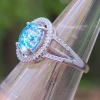 Close-up of an oval cremation ring in sterling silver featuring a bright blue memorial glass center stone surrounded by 52 cubic zirconia accents, shown on a clear display cone outdoors.