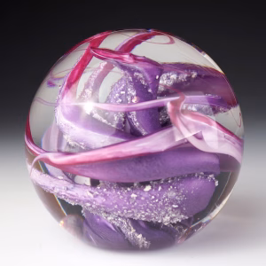 Ocean Memorial Cremation Paperweight featuring a hand-blown glass sphere with layered lavender and deep purple glass, accented by soft pink ribbons and delicate white inclusions suspended in clear glass.