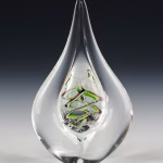 Teardrop memorial glass keepsake containing cremation ash, with a clear sculpted teardrop form and a green and purple spiral design suspended inside the glass, photographed upright against a dark studio background.