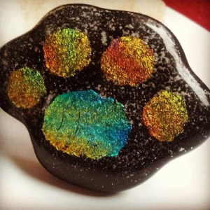 Handcrafted paw print cremation stone made with cremation ash, featuring a glossy black memorial glass base with iridescent multicolor paw pads and delicate ash inclusions throughout.
